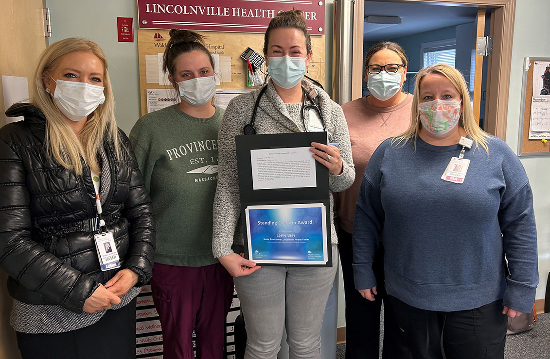 Five women, wearing masks and standing together at the Lincolnville Health Center. The center woman, Leslie Bray, is holding an award.