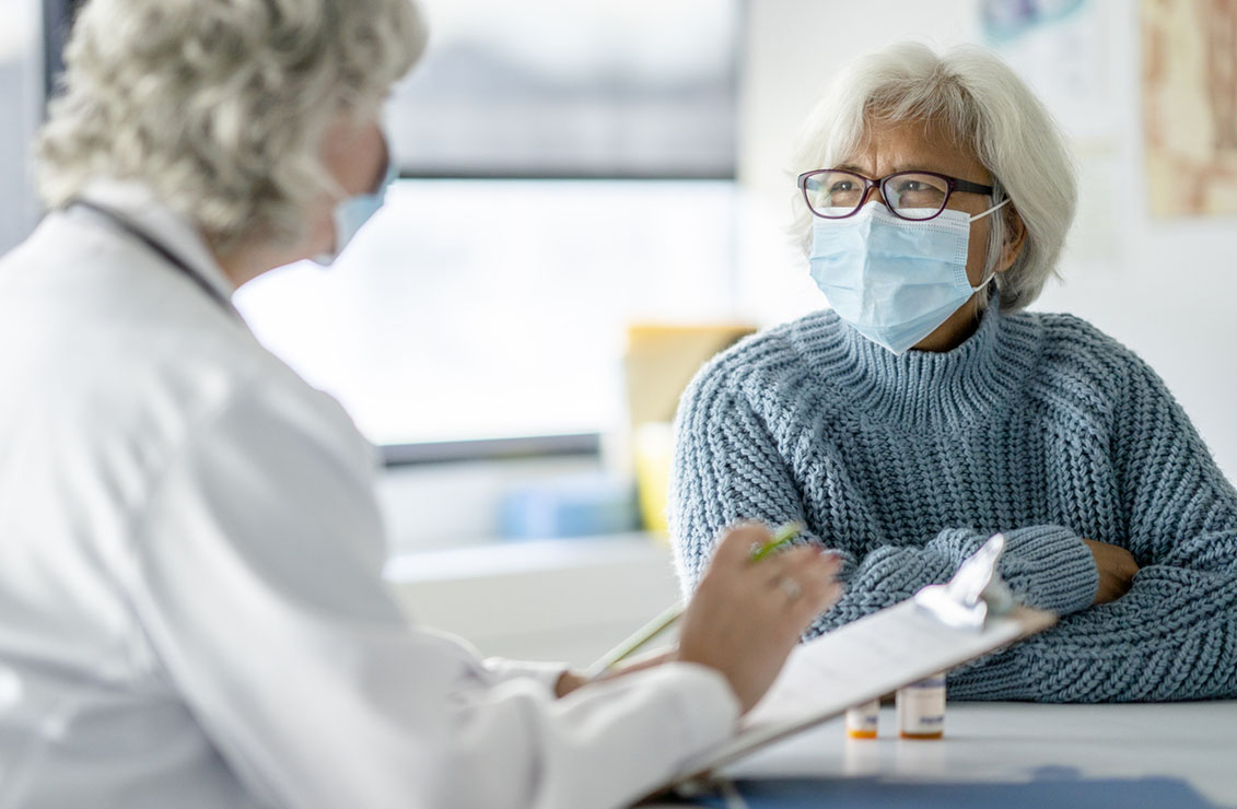 A female doctor holds out a clipboard during an appointment with her senior patient, as she asks her questions and collects her health history. The patient is dressed casually in a blue sweater and smiling contently. Both are wearing medical masks to protect them from the COVID-19 virus.